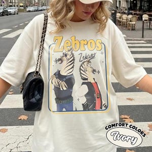 May include: Ivory t-shirt with a cartoon graphic of two zebras wearing outfits, the text "Zebros" and "Comfort Colors Ivory". The zebras are in a cartoon style, one in a vest and the other in a jacket.