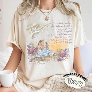 May include: An ivory t-shirt featuring a graphic illustration of a person with long blonde hair wearing a blue dress and boots, sitting in a field of purple and white flowers under a tree. The text reads, I remember I loved you, I knew it.