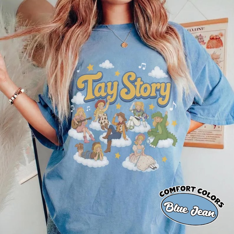 May include: A light blue t-shirt featuring the text Tay Story in yellow letters. The graphic includes multiple illustrations of a blonde character in various outfits, including a cowgirl, a dinosaur costume, and a dress, all positioned on white clouds.