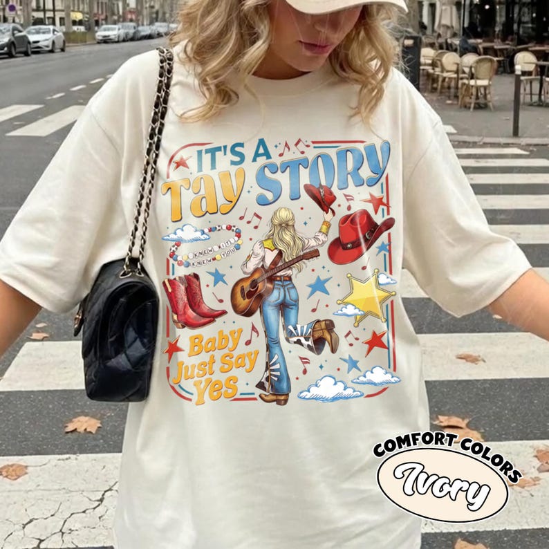 May include: An ivory t-shirt featuring a graphic print with the text IT'S A TAY STORY and Baby Just Say Yes. The design includes a blonde person playing a guitar, red cowboy boots, a red hat, a yellow star, clouds, and musical notes.