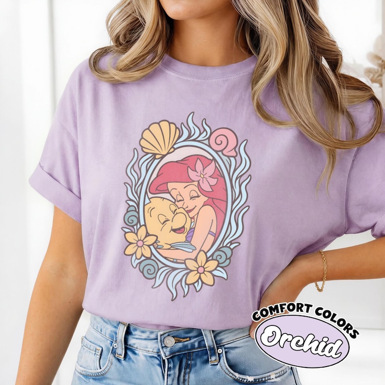 May include: A light purple short-sleeve t-shirt featuring a graphic print of a red-haired mermaid character hugging a yellow fish. The design is framed by an oval border of blue waves, yellow flowers, and seashells. The shirt has a crew neckline.