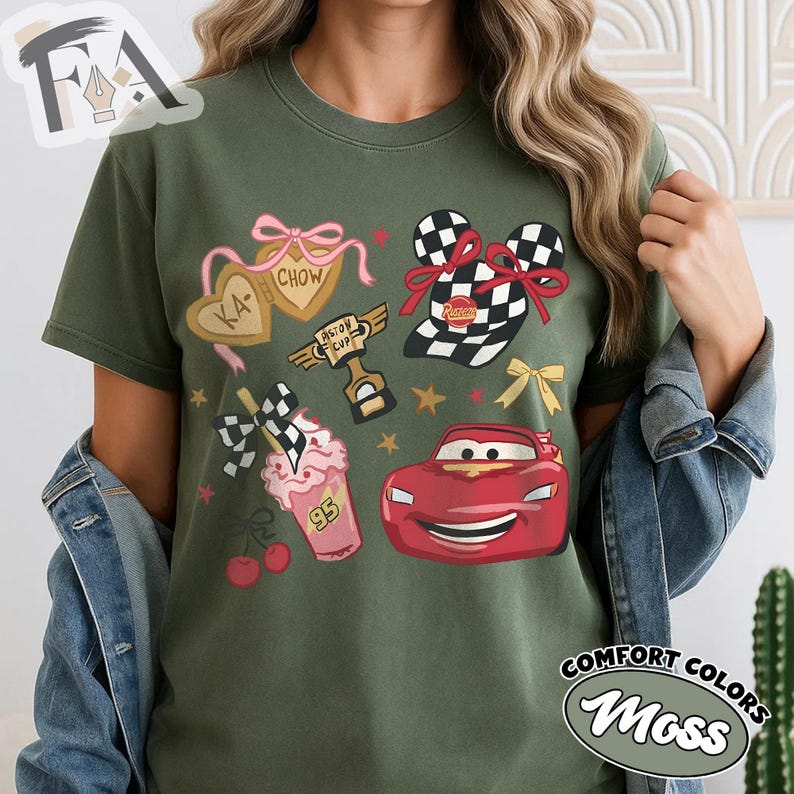 May include: Olive green t-shirt featuring cartoon graphics of a red race car, a checkered flag, and a milkshake. The shirt also includes the text "KA-CHOW" and "Piston Cup". The shirt is labeled "Comfort Colors Moss".