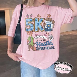 Puede incluir: Camiseta rosa claro con un dise&ntilde;o de dibujos animados. El dise&ntilde;o incluye el texto "5K" en huellas de patas azules, personajes de dibujos animados y la frase "...IT'S CALLED A Hustle, SWEETHEART." La camiseta tambi&eacute;n tiene las palabras "Comfort Colors Blossom".