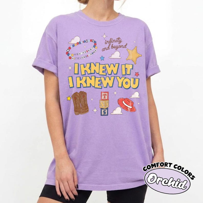 Pode incluir: Uma t-shirt roxo-claro com uma estampa gr&aacute;fica contendo o texto "I KNEW IT I KNEW YOU", "infinity and beyond" e "YOU'VE GOT A FRIEND IN ME". O design inclui ilustra&ccedil;&otilde;es de botas de cowboy, um chap&eacute;u vermelho, blocos de letras e estrelas.