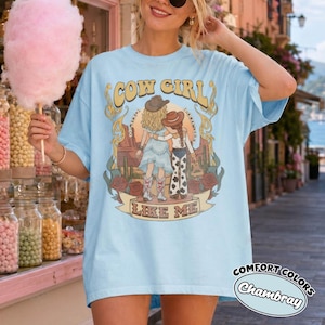 May include: A light blue t-shirt featuring a graphic print of two figures in cowboy hats standing in a desert landscape with cacti and roses. The text "COW GIRL" appears above the figures, and "LIKE ME" is printed on a banner below them.
