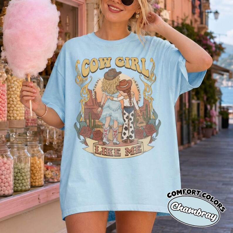 May include: A light blue t-shirt featuring a graphic print of two figures in cowboy hats standing in a desert landscape with cacti and roses. The text "COW GIRL" appears above the figures, and "LIKE ME" is printed on a banner below them.