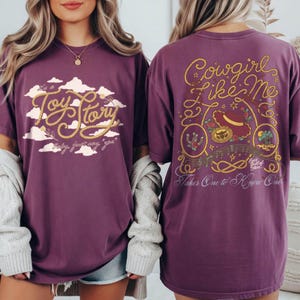 May include: A plum-colored t-shirt featuring a front graphic with the text Toy Story in rope-style lettering against white clouds. The back graphic displays the text Cowgirl Like Me with rope borders, a cowboy hat, and musical notes.