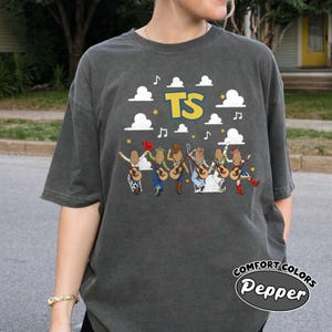 Może przedstawiać: Ciemnoszary t-shirt z kr&oacute;tkim rękawem z nadrukiem graficznym przedstawiającym sześć postaci z gitarami, białymi chmurami, ż&oacute;łtymi gwiazdami, nutami i ż&oacute;łtym napisem TS. Koszulka ma okrągły dekolt i luźny kr&oacute;j.