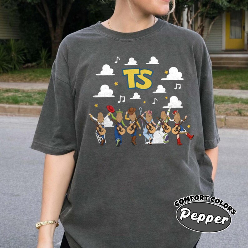 Może przedstawiać: Ciemnoszary t-shirt z kr&oacute;tkim rękawem z nadrukiem graficznym przedstawiającym sześć postaci z gitarami, białymi chmurami, ż&oacute;łtymi gwiazdami, nutami i ż&oacute;łtym napisem TS. Koszulka ma okrągły dekolt i luźny kr&oacute;j.