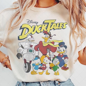 Puede incluir: Camiseta color crema con un gráfico de la caricatura de Disney "DuckTales". El diseño incluye el logotipo del programa y varios personajes, incluyendo Scrooge McDuck y sus sobrinos. El gráfico es en tonos de rojo, azul, amarillo y blanco.