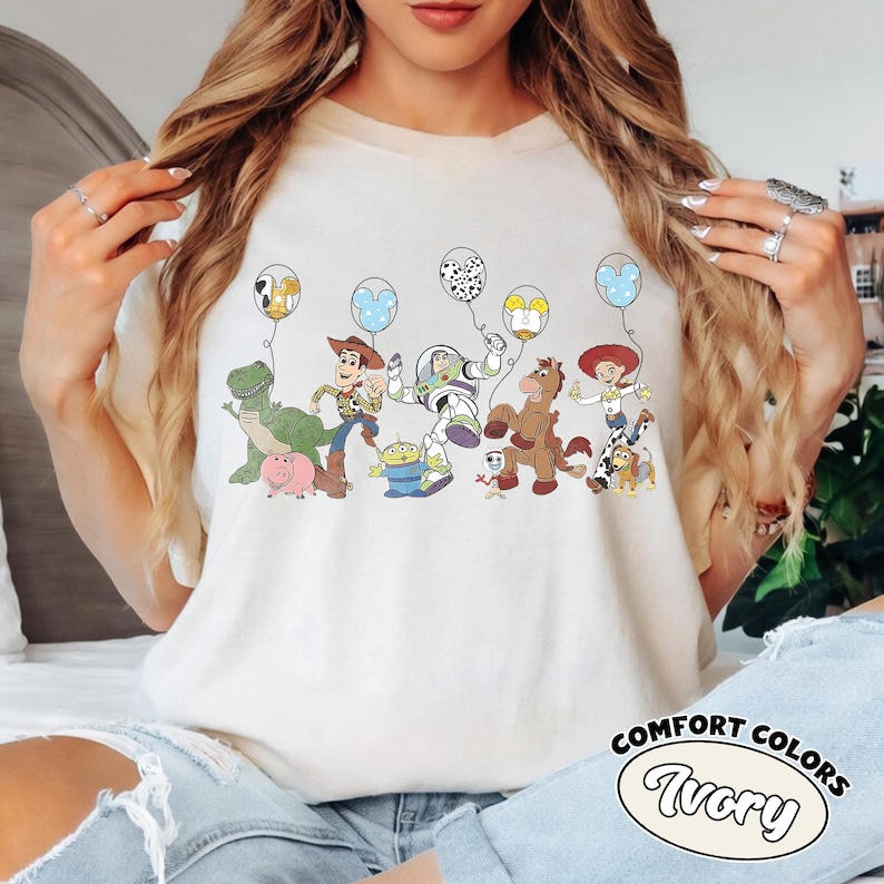 May include: Cream-coloured t-shirt with a vibrant graphic of Toy Story characters and balloons. The design includes Woody, Buzz Lightyear, and other characters from the animated film. The shirt has the words "Comfort Colors" and "Ivory" printed on it.