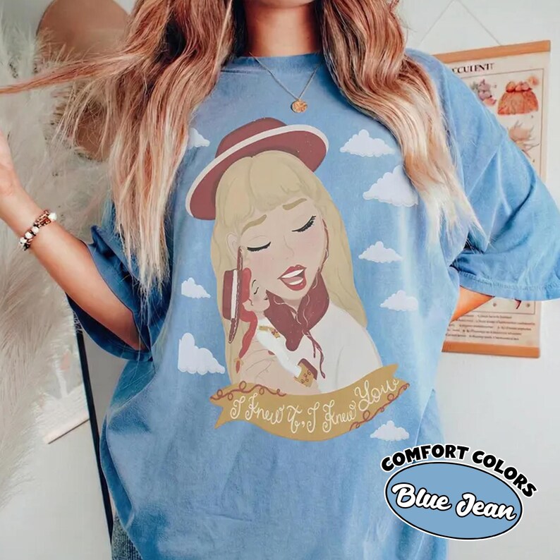Może przedstawiać: Jasnoniebieski t-shirt z grafiką przedstawiającą blond postać w bordowym kapeluszu, trzymającą małą lalkę. Wz&oacute;r zawiera białe chmury i złoty baner z brązowym napisem I Knew You, I Knew You.