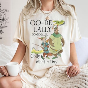 Puede incluir: Camiseta color crema con una ilustración de dibujos animados de Robin Hood y Little John. El texto dice "OO-DE-LALLY, OO-DE-LALLY, Golly, What a Day!". El diseño incluye detalles verdes, marrones y azules.