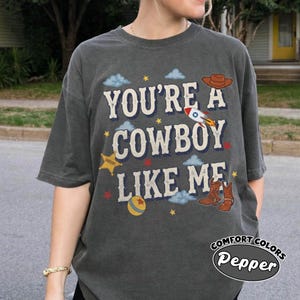 May include: A charcoal gray short-sleeve t-shirt featuring the text "YOU'RE A COWBOY LIKE ME" in a cream-colored font. The design includes illustrations of a brown cowboy hat, a rocket ship, a yellow sheriff star, a ball, and brown cowboy boots.
