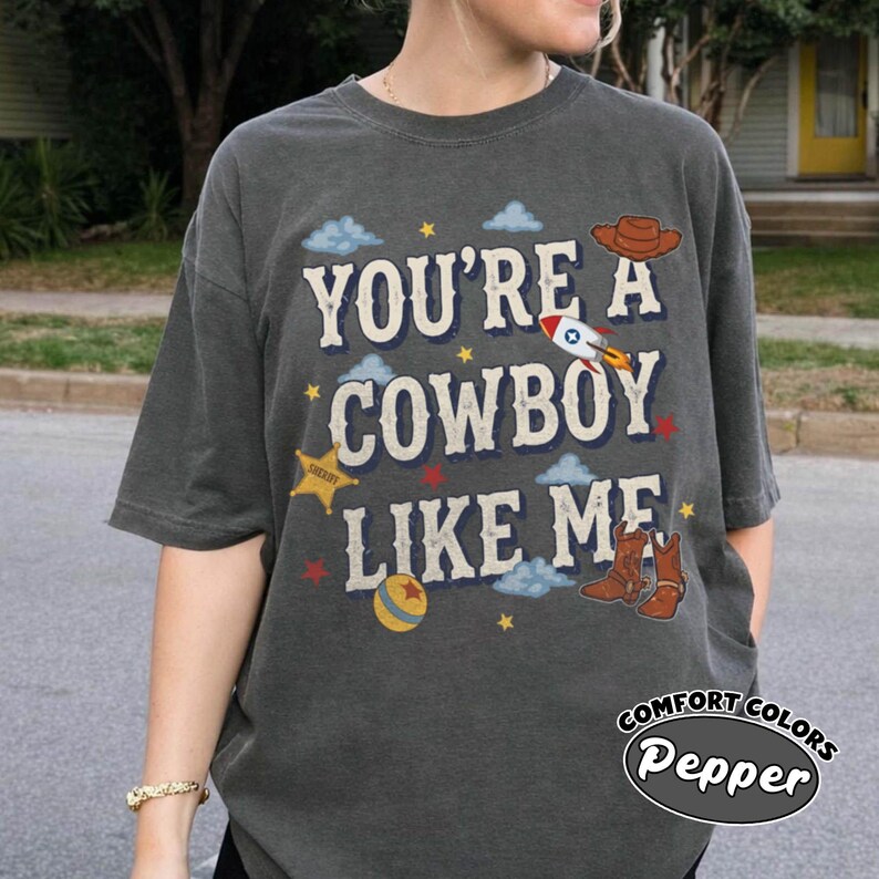 May include: A charcoal gray short-sleeve t-shirt featuring the text "YOU'RE A COWBOY LIKE ME" in a cream-colored font. The design includes illustrations of a brown cowboy hat, a rocket ship, a yellow sheriff star, a ball, and brown cowboy boots.