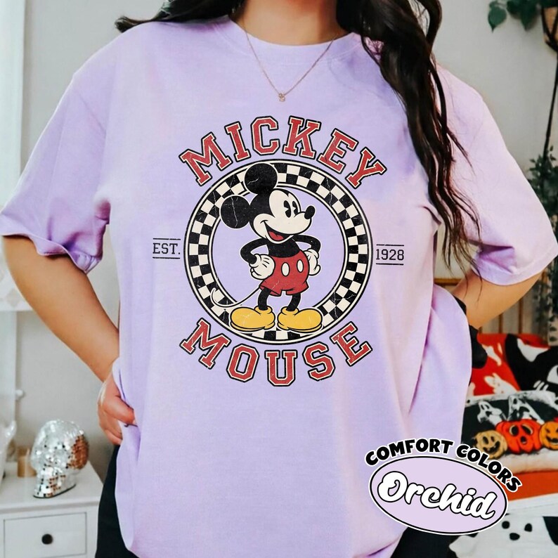 May include: A light purple t-shirt featuring a graphic of Mickey Mouse in a circular design with the words "Mickey Mouse" and "Est. 1928". The shirt also has the words "Comfort Colors" and "Orchid" printed on it.