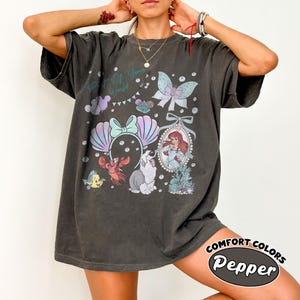 May include: A charcoal gray short-sleeve t-shirt featuring a graphic print with the text Part of Your World. The design includes a mermaid, a crab, a fish, a dog, a bow, and a butterfly in pastel colors, arranged with bubbles and sea elements.