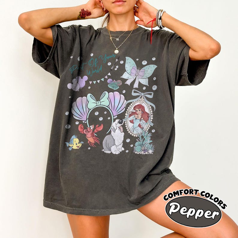 May include: A charcoal gray short-sleeve t-shirt featuring a graphic print with the text Part of Your World. The design includes a mermaid, a crab, a fish, a dog, a bow, and a butterfly in pastel colors, arranged with bubbles and sea elements.