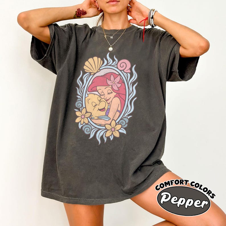 May include: A charcoal gray short-sleeve t-shirt featuring a graphic print of a cartoon mermaid with red hair hugging a yellow fish. The design is framed by an oval border of blue waves, seashells, and flowers. The shirt has a crew neckline.