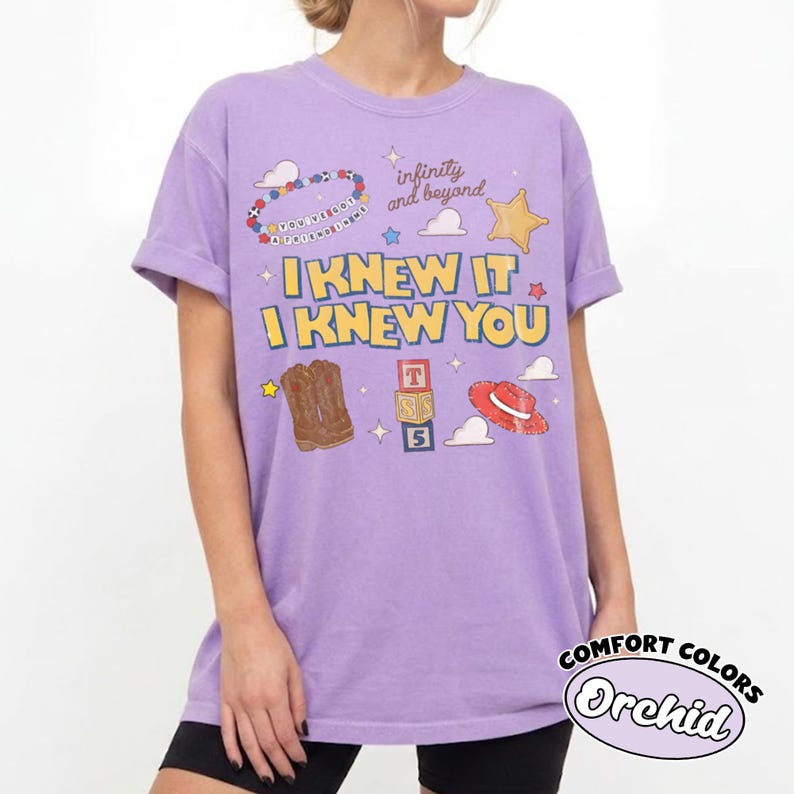 May include: A light purple t-shirt featuring the text I KNEW IT I KNEW YOU in yellow letters. The graphic includes a beaded bracelet, a yellow star, brown cowboy boots, a stack of alphabet blocks, a red hat, and the text infinity and beyond.