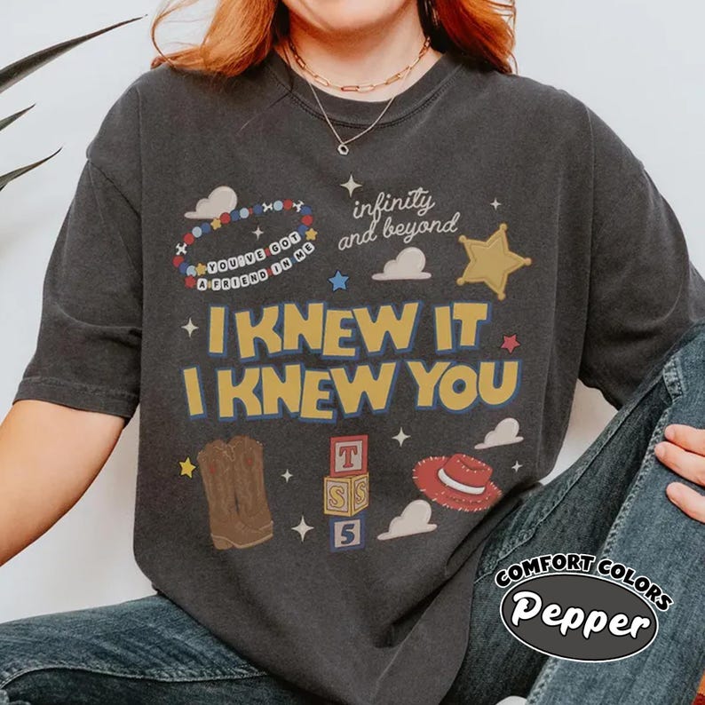 May include: A charcoal gray short-sleeve t-shirt featuring a graphic print with the text "I KNEW IT I KNEW YOU", "infinity and beyond", and "YOU'VE GOT A FRIEND IN ME". The design includes illustrations of a beaded bracelet, a star, clouds, cowboy boots, alphabet blocks, and a red cowboy hat.
