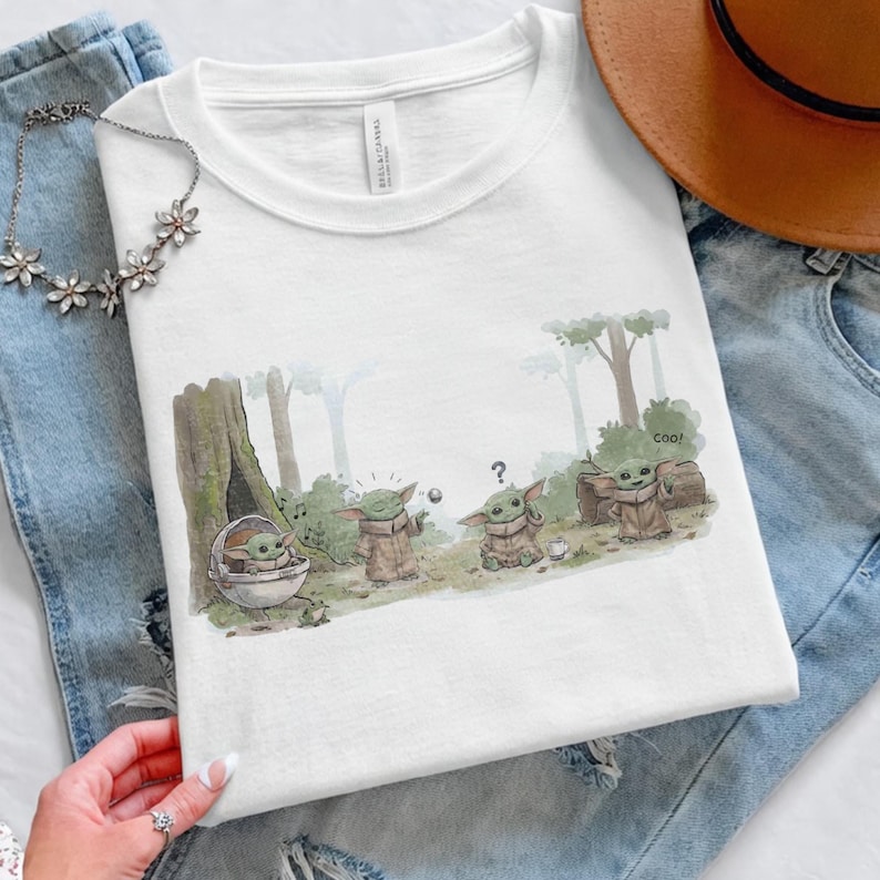 Puede incluir: Camiseta blanca de cuello redondo con una ilustraci&oacute;n de dibujos animados de Grogu de The Mandalorian. El dise&ntilde;o muestra a Grogu en varias poses, con la palabra "cool" y un signo de interrogaci&oacute;n. La camiseta est&aacute; doblada sobre unos vaqueros.