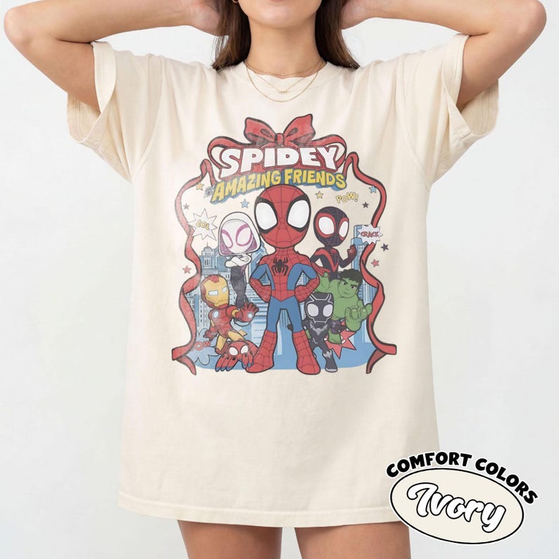 May include: Ivory-colored t-shirt featuring a graphic of Spidey and his Amazing Friends. The design includes Spider-Man, Iron Man, and other characters. The shirt has the words "Spidey Amazing Friends" in a red ribbon design. The shirt is labeled "Comfort Colors Ivory".