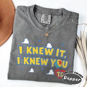May include: A charcoal gray t-shirt featuring the yellow text I KNEW IT, I KNEW YOU. The design includes white cloud graphics, small blue stars, a colorful ball with a red star, and a red heart with the letters TS. The shirt has a crew neckline.