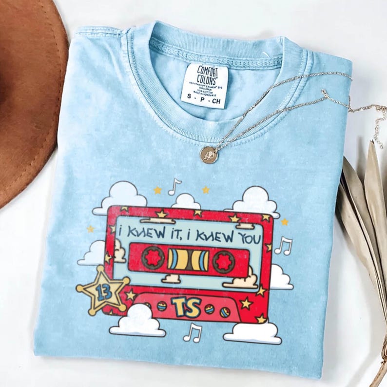 May include: A light blue t-shirt featuring a graphic of a red cassette tape with the text "I KNEW IT, I KNEW YOU" and "TS". The design includes white clouds, yellow stars, and musical notes. A silver chain necklace rests on the neckline.