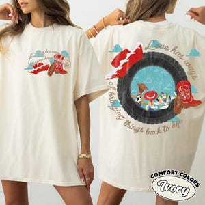 May include: An ivory short-sleeve t-shirt featuring a graphic print of a tire, a red cowboy boot, and characters from Toy Story. The text "Love has ways of bringing things back to life" is printed in a cursive font around the tire graphic.