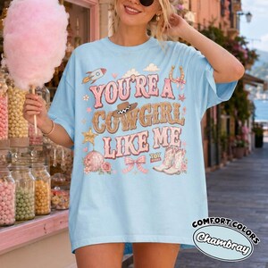 May include: A light blue t-shirt featuring a graphic print with the text "YOU'RE A COWGIRL LIKE ME" in pink and brown letters. The design includes a cowboy hat, a rocket, a horseshoe, a disco ball, a bow, and cowboy boots in a western style.