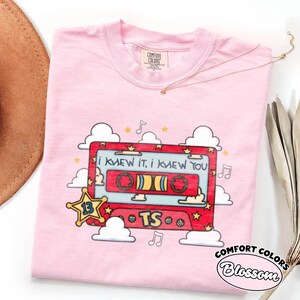 May include: A light pink t-shirt featuring a graphic of a red cassette tape with the text "I KNEW IT, I KNEW YOU" and "TS". The design includes a yellow star with the number 13, white clouds, and musical notes. The shirt has a Comfort Colors label.