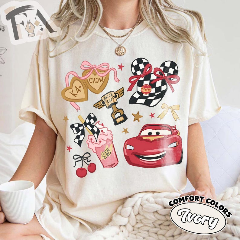 May include: Ivory-colored t-shirt featuring cartoon graphics of a red race car, checkered flags, and heart-shaped cookies. The shirt also includes a milkshake, cherries, and the text "Piston Cup". The shirt is labeled "Comfort Colors Ivory".