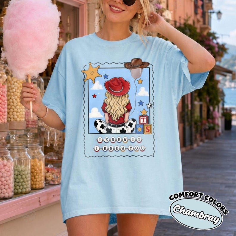 May include: A light blue t-shirt featuring a graphic of a blonde person wearing a red cowboy hat, sitting with their back to the viewer. The design includes a yellow star, a balloon, alphabet blocks, and the text I KNEW IT I KNEW YOU.
