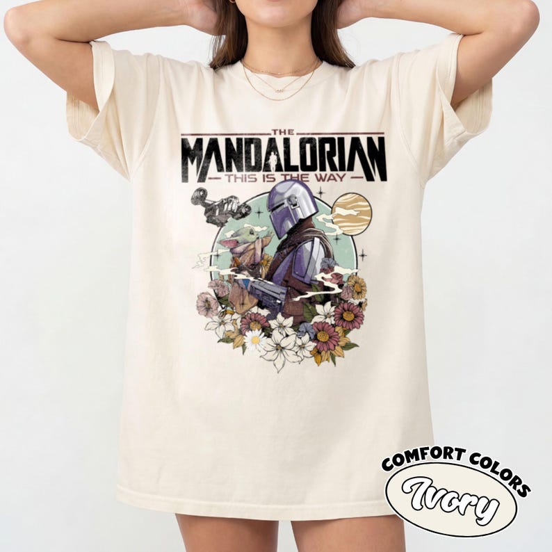 Op de afbeelding: Een ivoorkleurig T-shirt met korte mouwen en een grafische print van The Mandalorian en Grogu. De tekst luidt THE MANDALORIAN THIS IS THE WAY. Het ontwerp bevat een cirkelvormige achtergrond met een planeet, een ruimteschip en bloemen.