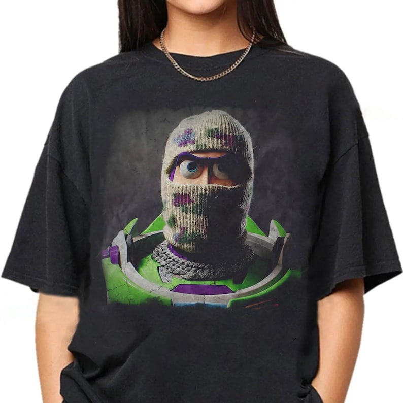 K&ouml;nnte beinhalten: Ein schwarzes T-Shirt mit kurzen &Auml;rmeln und einem Grafikdruck einer Figur, die eine beige Camouflage-Sturmhaube und eine silberne Kette tr&auml;gt. Die Figur hat sichtbare blaue Augen und steckt in einer gr&uuml;nen und violetten Weltraumr&uuml;stung.