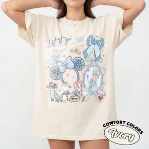 May include: An ivory t-shirt featuring a graphic print with the text Let it go. The design includes a blue bow, snowflake-patterned ears, a mirror with a character illustration, a lizard, a snowman, popcorn, a drink, and a ticket labeled Magic.