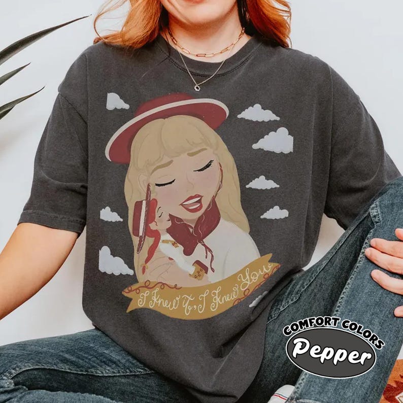 Może przedstawiać: Ciemnoszary t-shirt z grafiką przedstawiającą blond postać w czerwonym kapeluszu, trzymającą małą lalkę. Wz&oacute;r zawiera białe chmury oraz złotą wstęgę z napisem "I Knew It, I Knew You" wykonanym ozdobną czcionką przypominającą pismo odręczne.