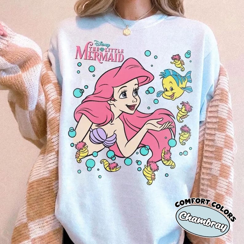 Puede incluir: Una camiseta azul claro con un gr&aacute;fico de Ariel de La Sirenita con largo cabello rosa, un top de concha morado y un pez amarillo. El dise&ntilde;o incluye varios caballitos de mar amarillos peque&ntilde;os y burbujas color verde azulado.