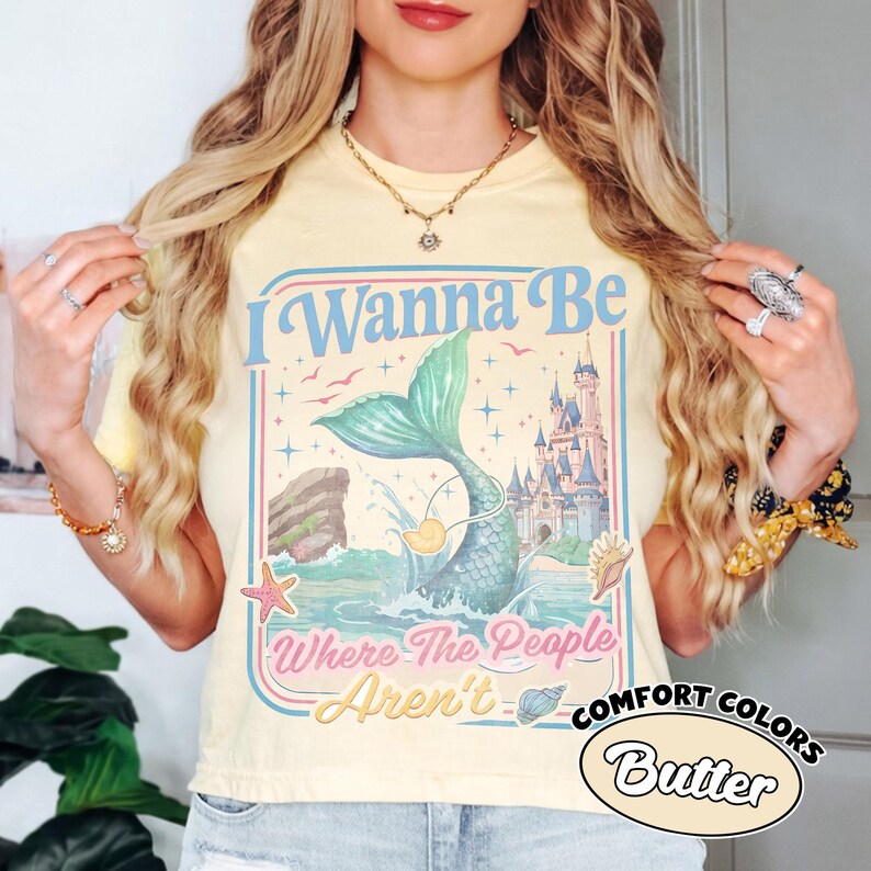 May include: Butter-colored t-shirt with a mermaid graphic and the text "I Wanna Be Where The People Aren't." The design includes a mermaid tail, castle, and seashells. The shirt is part of the Comfort Colors collection.