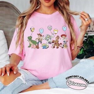 May include: A pink t-shirt with a cartoon illustration of Toy Story characters, each with a balloon. The shirt has the words "COMFORT COLORS Blossom" in a circular design.