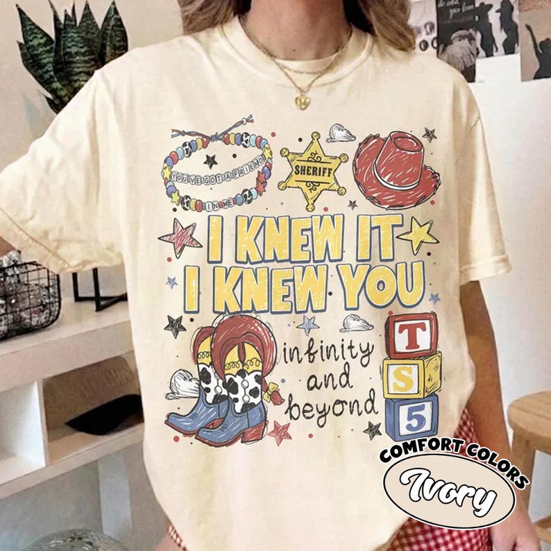 May include: An ivory short-sleeve t-shirt featuring a graphic print with the text I KNEW IT I KNEW YOU and infinity and beyond. The design includes a sheriff badge, a red cowboy hat, a beaded bracelet, cowboy boots, and stacked alphabet blocks.