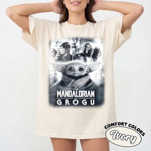 May include: Ivory-coloured t-shirt featuring a graphic from "The Mandalorian" series. The design includes Grogu, the Mandalorian, and other characters. The text "THE MANDALORIAN AND GROGU" is printed below the image. The shirt is labelled "COMFORT COLORS Ivory".