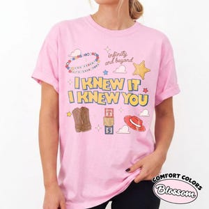 May include: A light pink t-shirt featuring the text I KNEW IT I KNEW YOU in yellow with blue outlines. Graphics include a beaded bracelet, a star, clouds, brown cowboy boots, alphabet blocks with the letters T, S, 5, and a red cowboy hat.