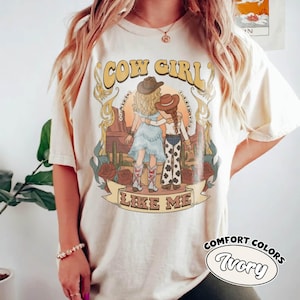 May include: An ivory short-sleeve t-shirt featuring a graphic print of two figures in cowboy hats standing in a desert landscape with cacti and roses. The text COW GIRL LIKE ME is printed in a decorative font. The design includes muted earth tones.