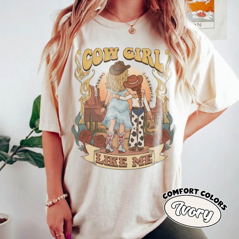 May include: An ivory short-sleeve t-shirt featuring a graphic print of two figures in cowboy hats standing in a desert landscape with cacti and roses. The text COW GIRL LIKE ME is printed in a decorative font. The design includes muted earth tones.