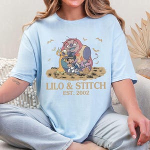 K&ouml;nnte beinhalten: Hellblaues T-Shirt mit einer Grafik von Lilo & Stitch-Charakteren. Das Design umfasst Stitch, Lilo und andere Charaktere mit dem Text "LILO & STITCH EST. 2002" in Gold. Flederm&auml;use sind ebenfalls im Design enthalten.