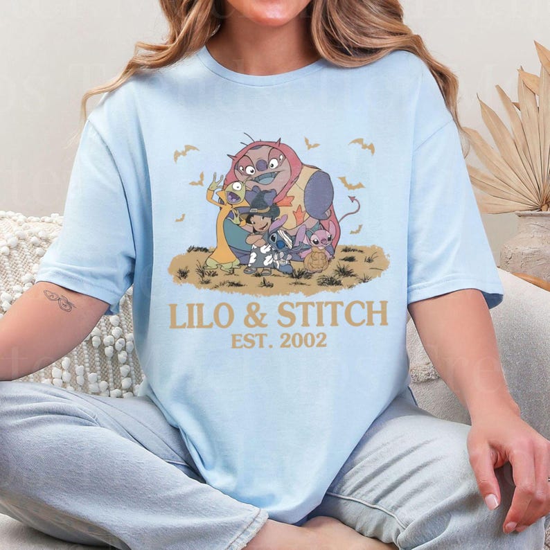 K&ouml;nnte beinhalten: Hellblaues T-Shirt mit einer Grafik von Lilo & Stitch-Charakteren. Das Design umfasst Stitch, Lilo und andere Charaktere mit dem Text "LILO & STITCH EST. 2002" in Gold. Flederm&auml;use sind ebenfalls im Design enthalten.