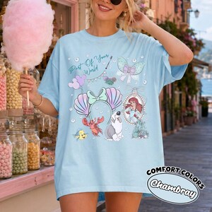 May include: A light blue t-shirt featuring a graphic print with the text Part Of Your World, a mermaid character, a crab, a fish, a dog, a seashell headband, a bow, and bubbles. The shirt has short sleeves and a crew neckline.