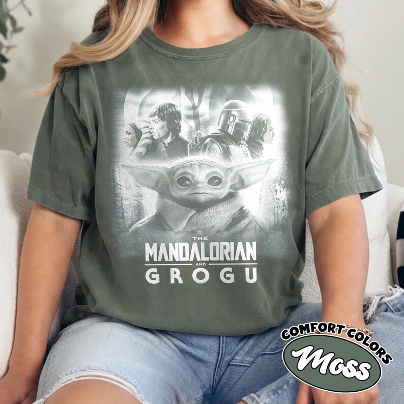 May include: Moss green t-shirt featuring a graphic of Grogu and The Mandalorian. The design includes characters from the Star Wars series, with the text "THE MANDALORIAN and GROGU" in white. The shirt is a Comfort Colors brand.